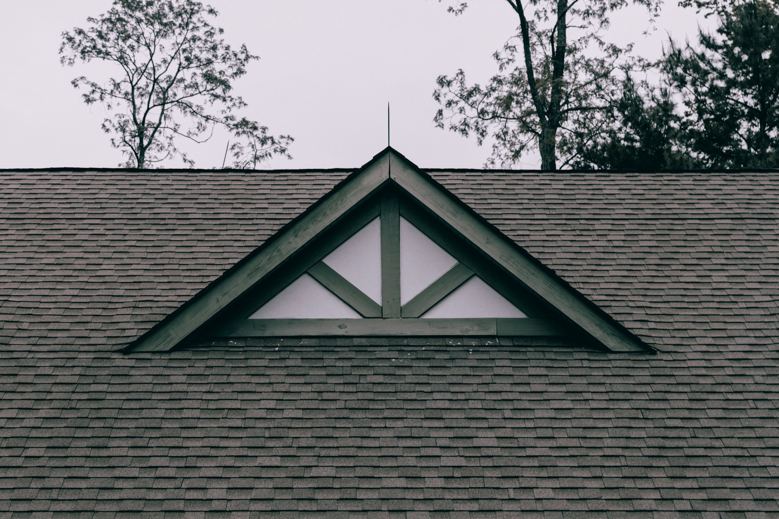The roof of a house with brown shingles and a triangular gable with decorative trim, with trees and a gray sky in the background.