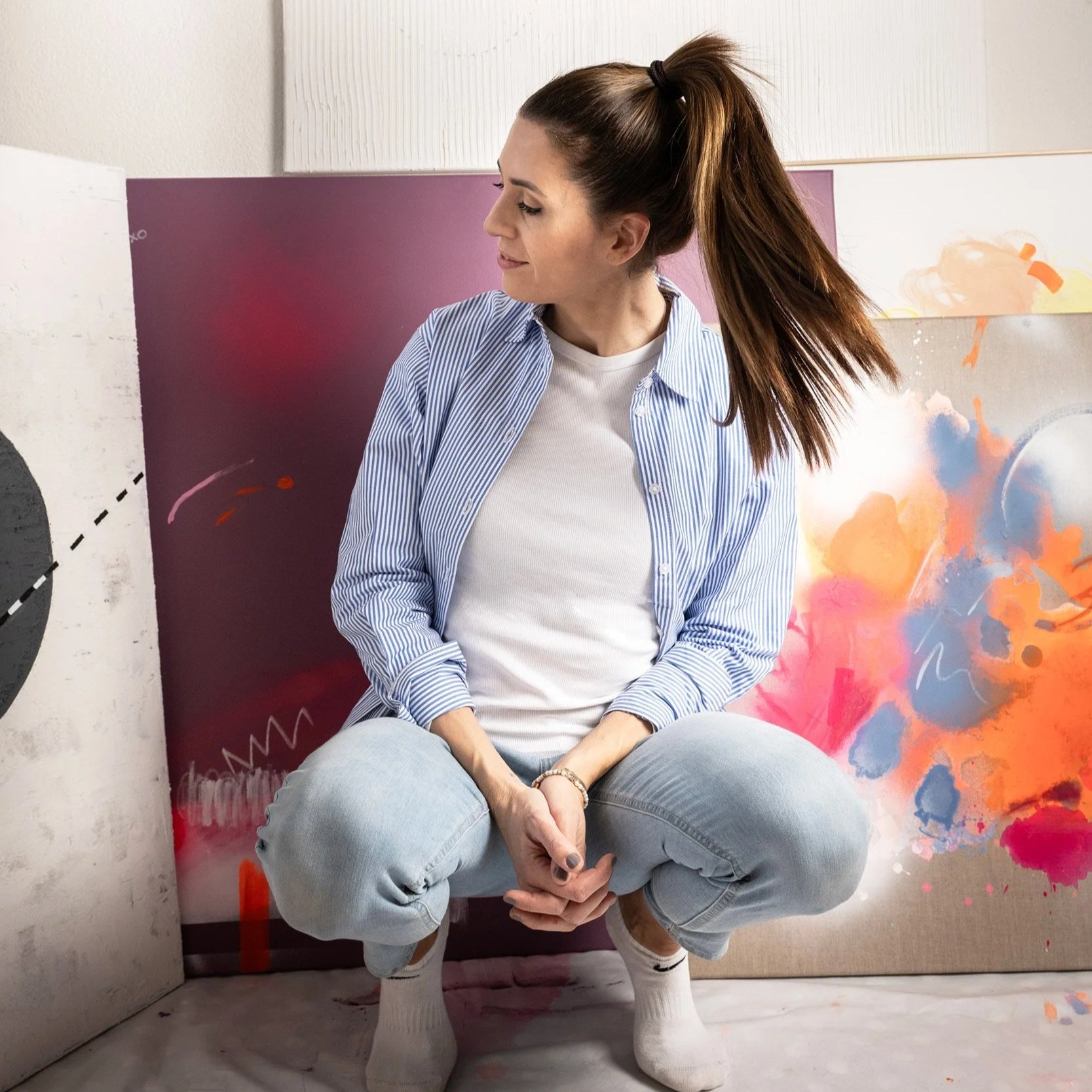 A woman sitting on the floor in front of colorful abstract paintings, wearing a white shirt, blue striped shirt, and light jeans.