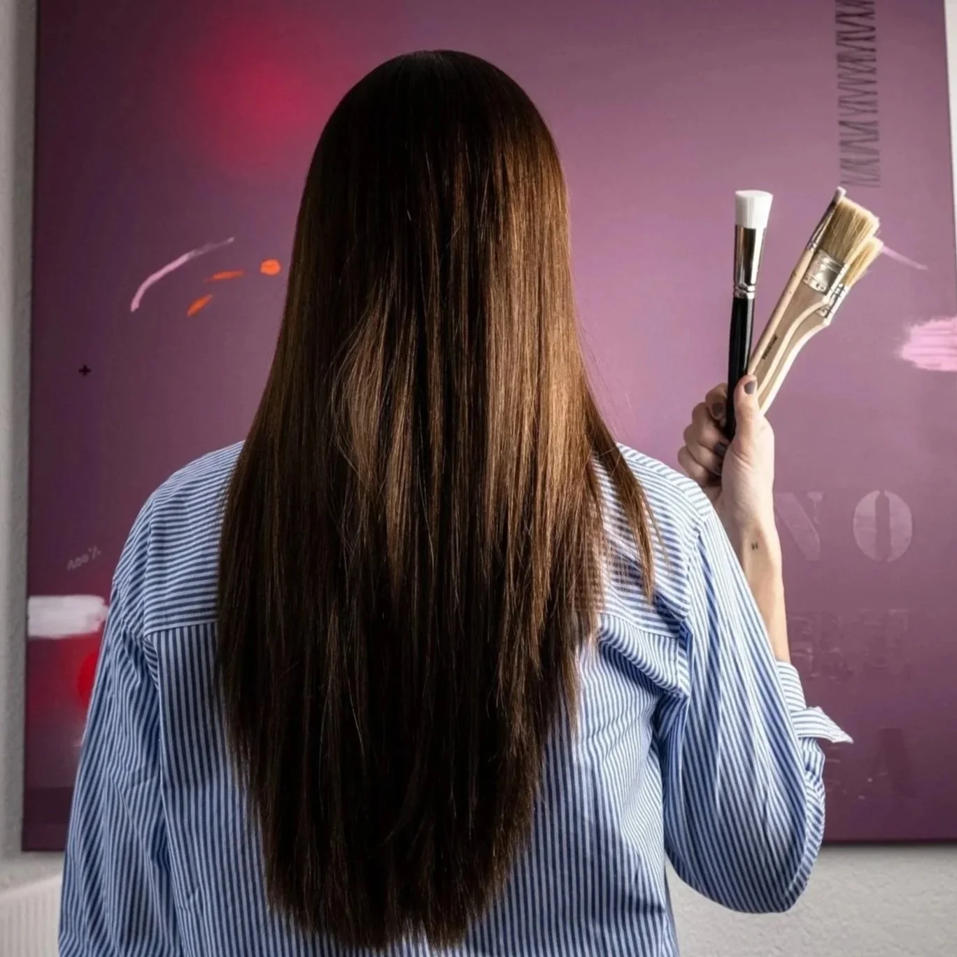 A woman with long brown hair holding paintbrushes, standing in front of a purple background with artistic markings.