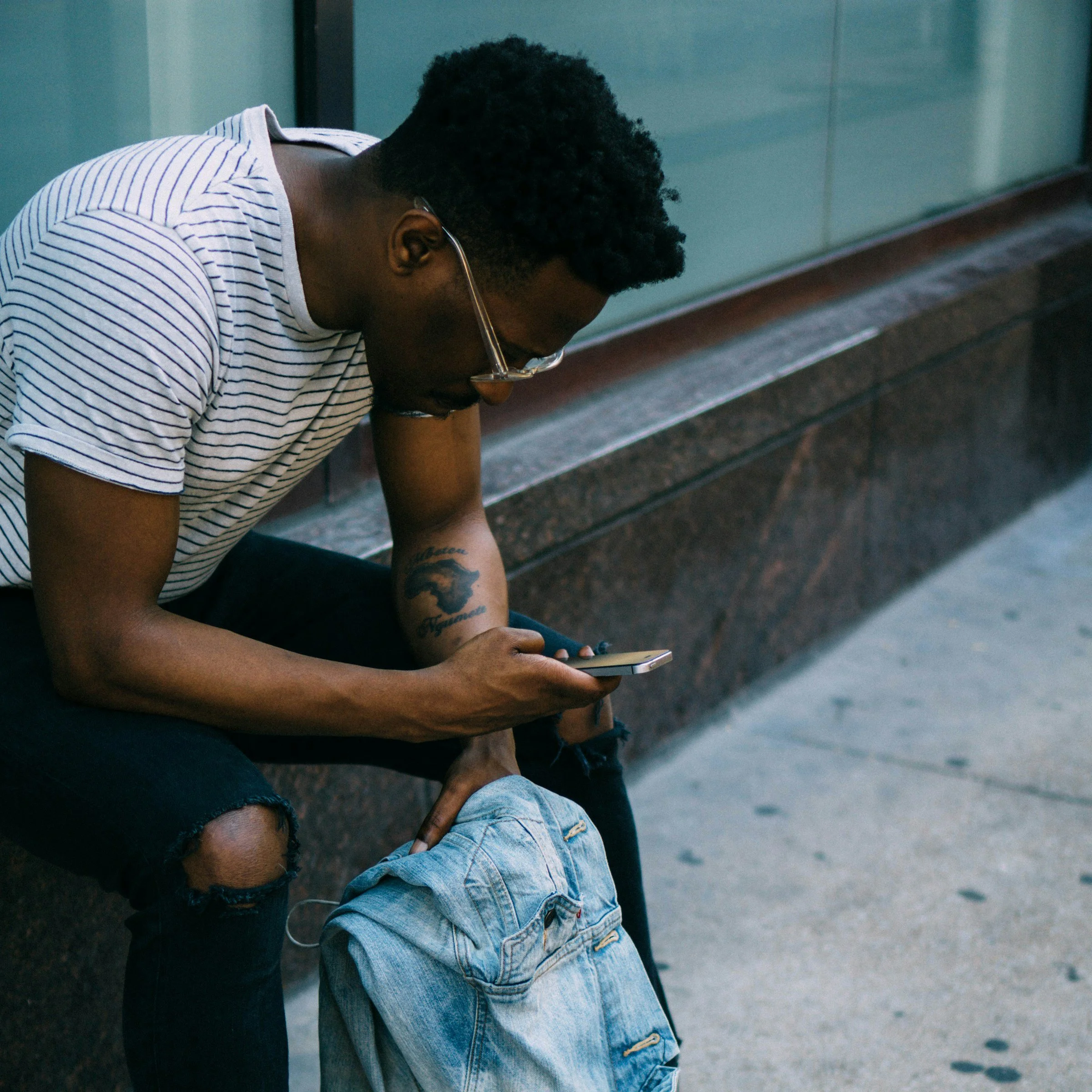 Young man with curly black hair, glasses, and a tattoo on his right arm, sitting on a bench and looking at his phone. He is wearing a striped t-shirt and ripped black jeans, with a pair of denim shorts and a dark backpack on the ground nearby.