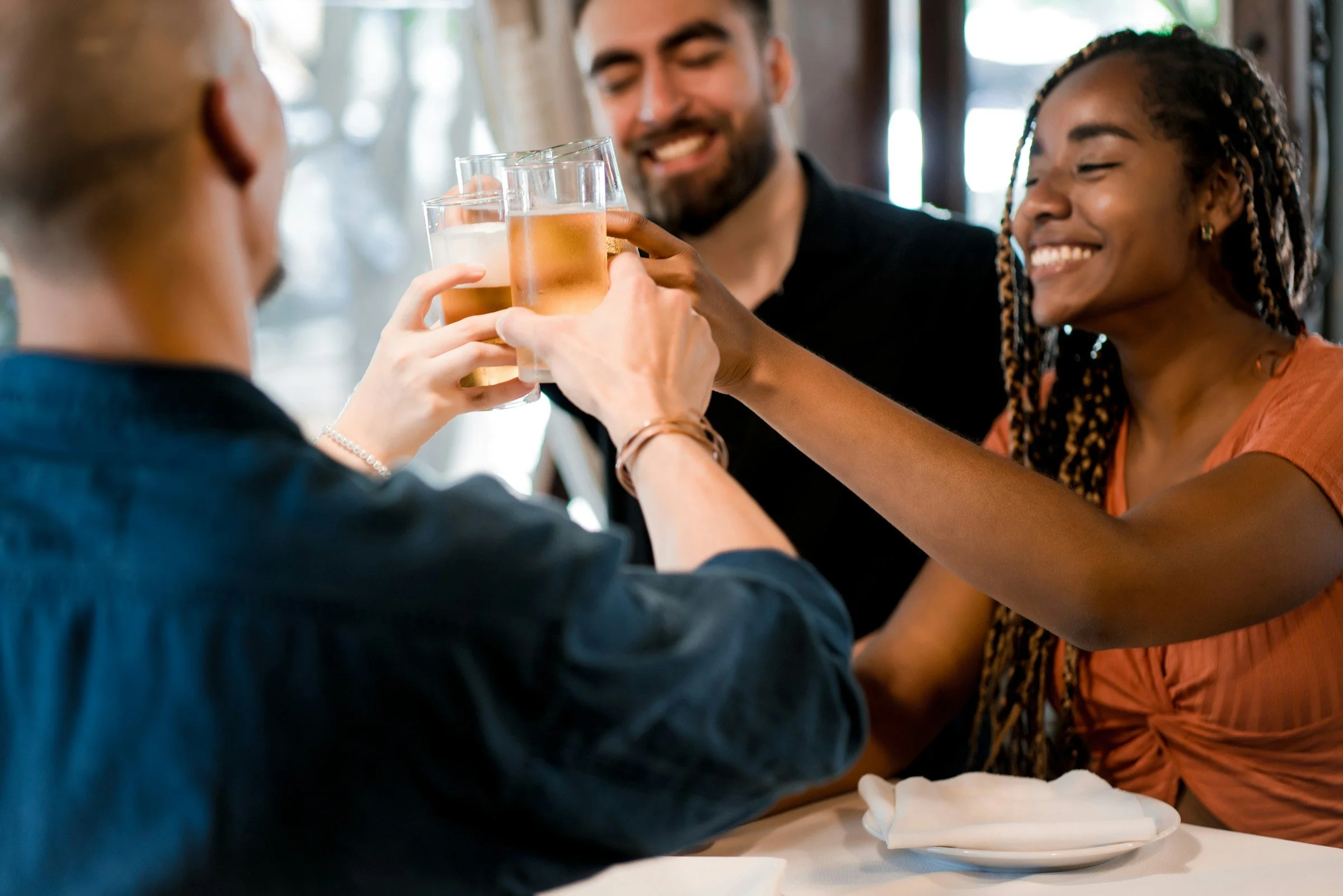 Three friends cheer with glasses of beer at a restaurant table, smiling and enjoying their time together.