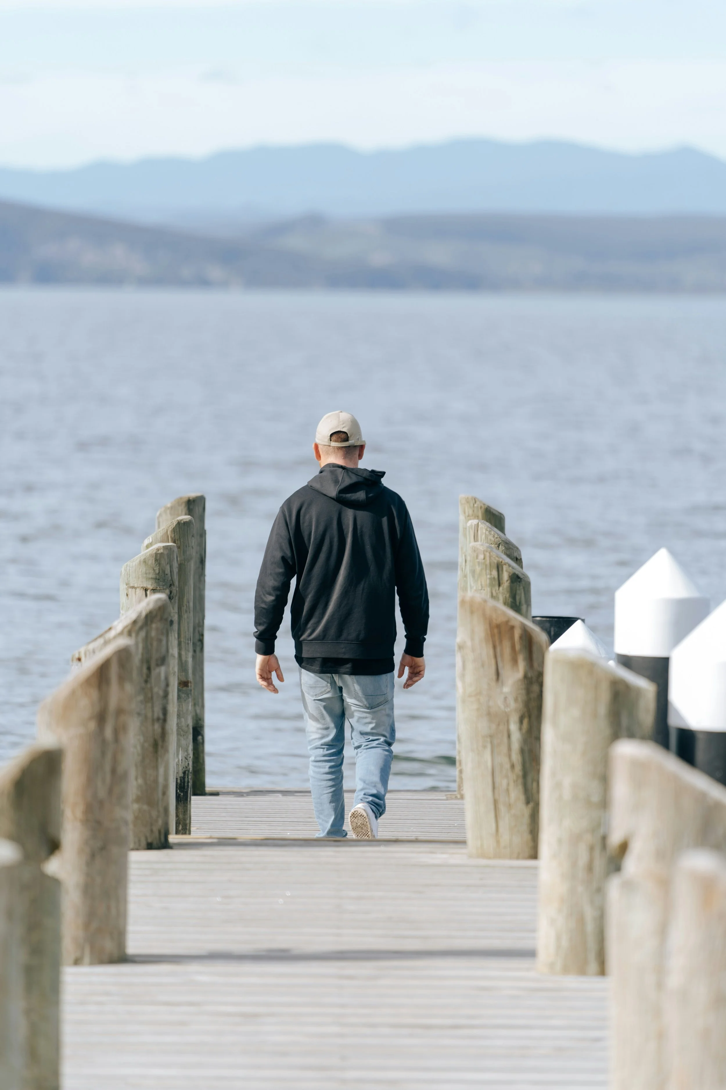 A man walking alone on a wooden dock towards a lake with mountains in the background.