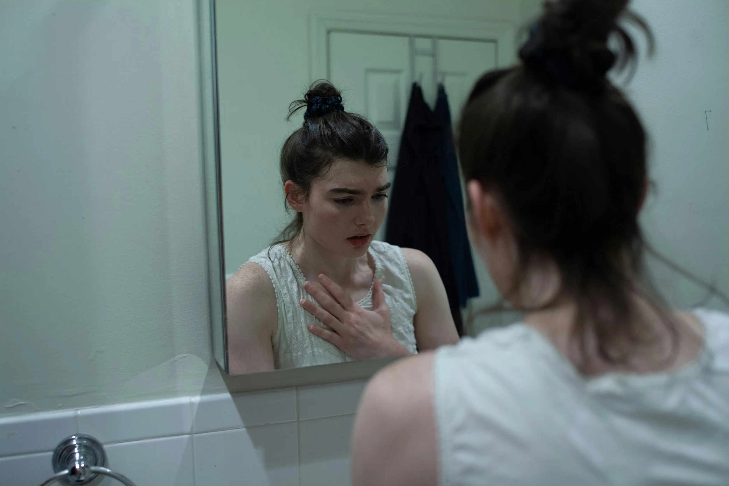 A young woman with dark hair in a messy bun looking at herself in a bathroom mirror, appearing worried or distressed, with her hand on her chest.