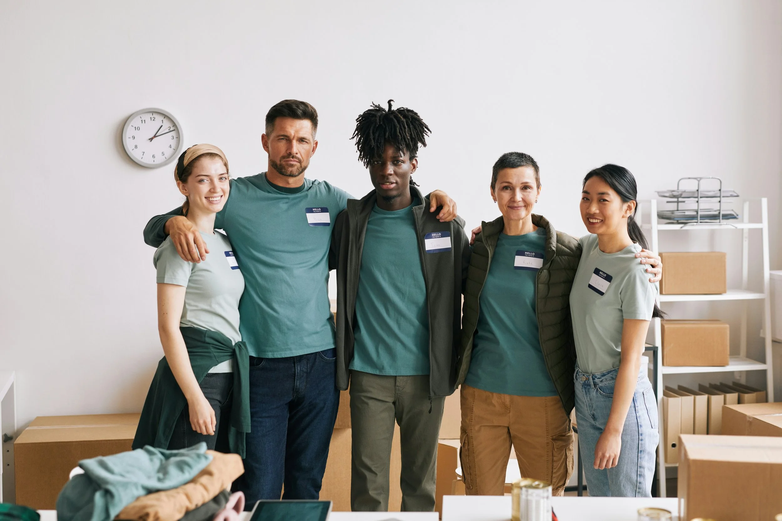 Group of five diverse volunteers in a room, wearing matching teal shirts and name tags, standing together with arms around each other, smiling.