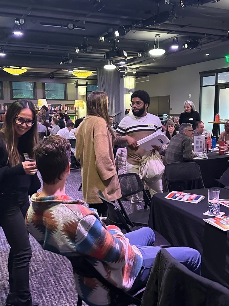 A group of people gathered in a modern, well-lit room with bookshelves and large windows, engaged in conversation and activities during a social event or conference.