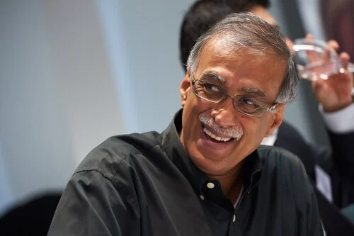 Man with glasses smiling and wearing a black button-up shirt at an indoor event.