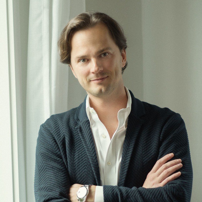 A man with light brown hair and a slight smile wearing a white shirt and black cardigan, standing with arms crossed in front of a window with white curtains.