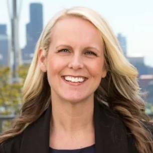 Portrait of a smiling woman with blonde hair outdoors, city skyline in the background.