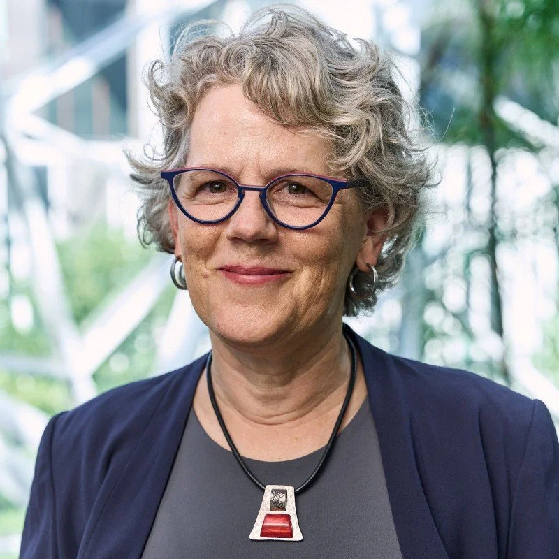 A middle-aged woman with curly gray hair and glasses, smiling outdoors with greenery and architectural structures in the background.