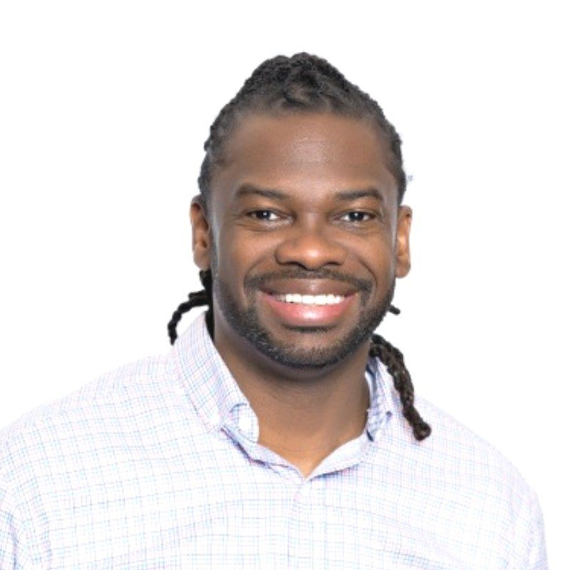 Portrait of a smiling man with dark hair and facial hair, wearing a light-colored checkered shirt, against a white background.