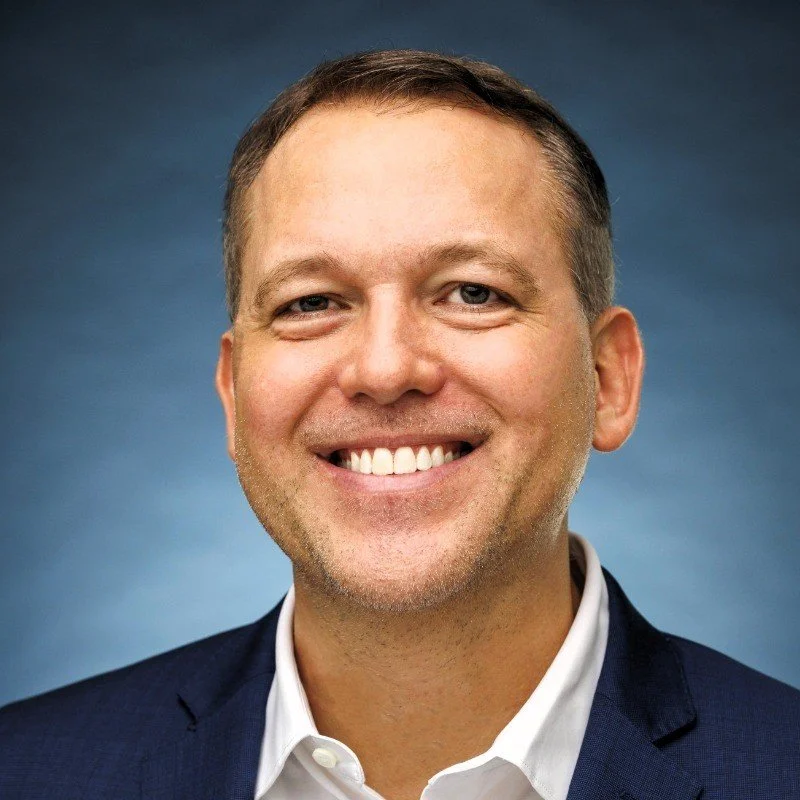Professional headshot of a smiling man wearing a dark blazer and a white collared shirt, with a blue gradient background.