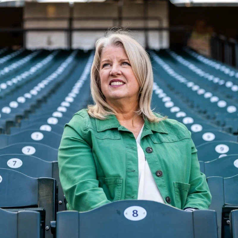 A woman with blonde hair wearing a green jacket sitting in an empty outdoor stadium with numbered seats.