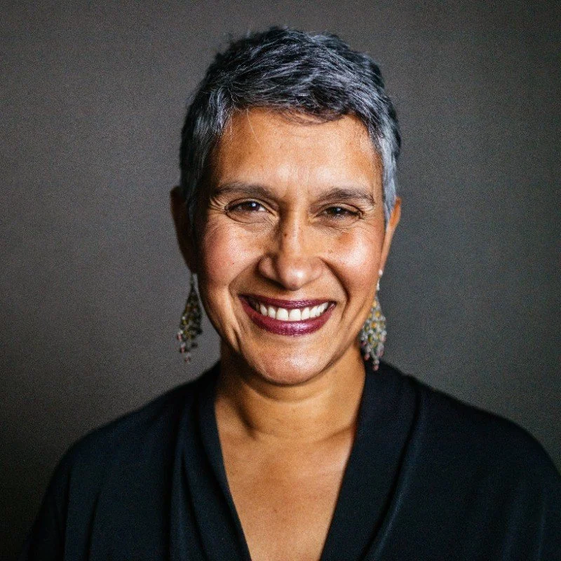 A woman with short gray hair smiling, wearing earrings and a dark top, against a dark background.