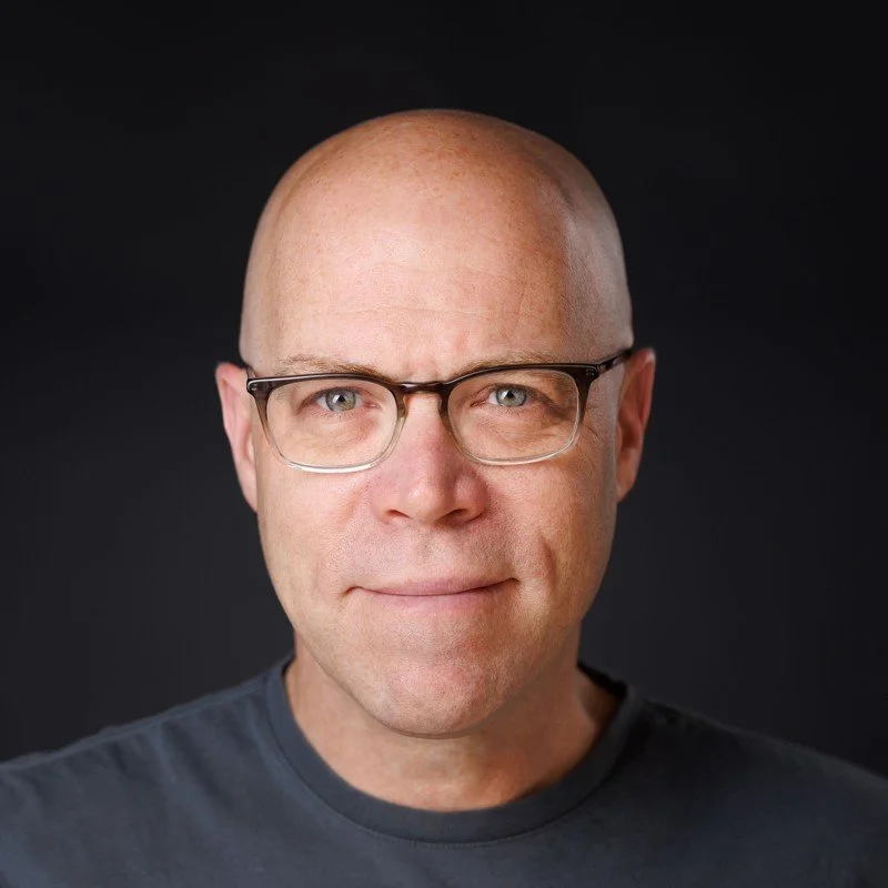 A man with a bald head, glasses, and a slight smile, wearing a dark gray shirt against a black background.