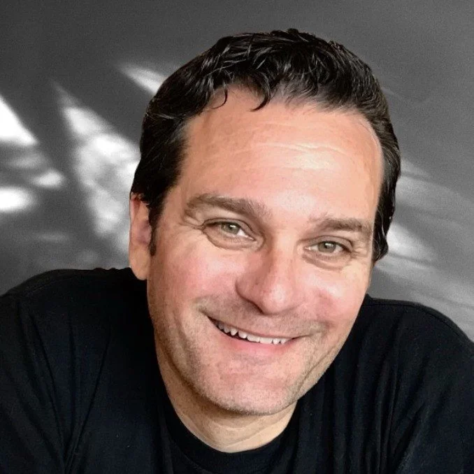 Smiling man with short dark hair, wearing a black shirt, against a gray background.