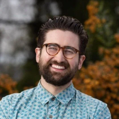 A smiling man with dark hair and a beard, wearing glasses and a blue patterned shirt, standing outdoors with autumn foliage in the background.