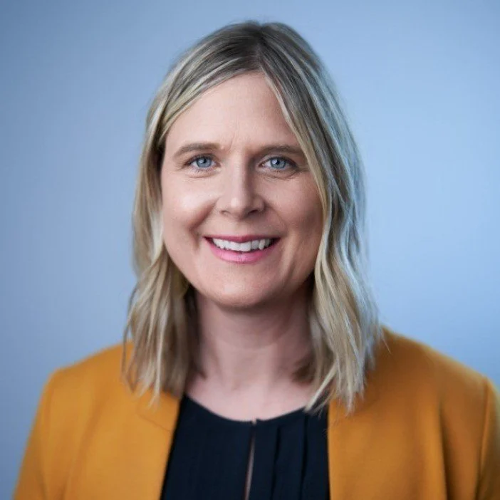 A woman with blonde hair and blue eyes smiling, wearing a black top and mustard-colored blazer, against a light blue background.
