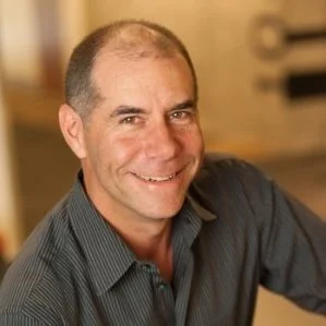 A smiling man with short hair, wearing a dark collared shirt, looking at the camera in an indoor setting.