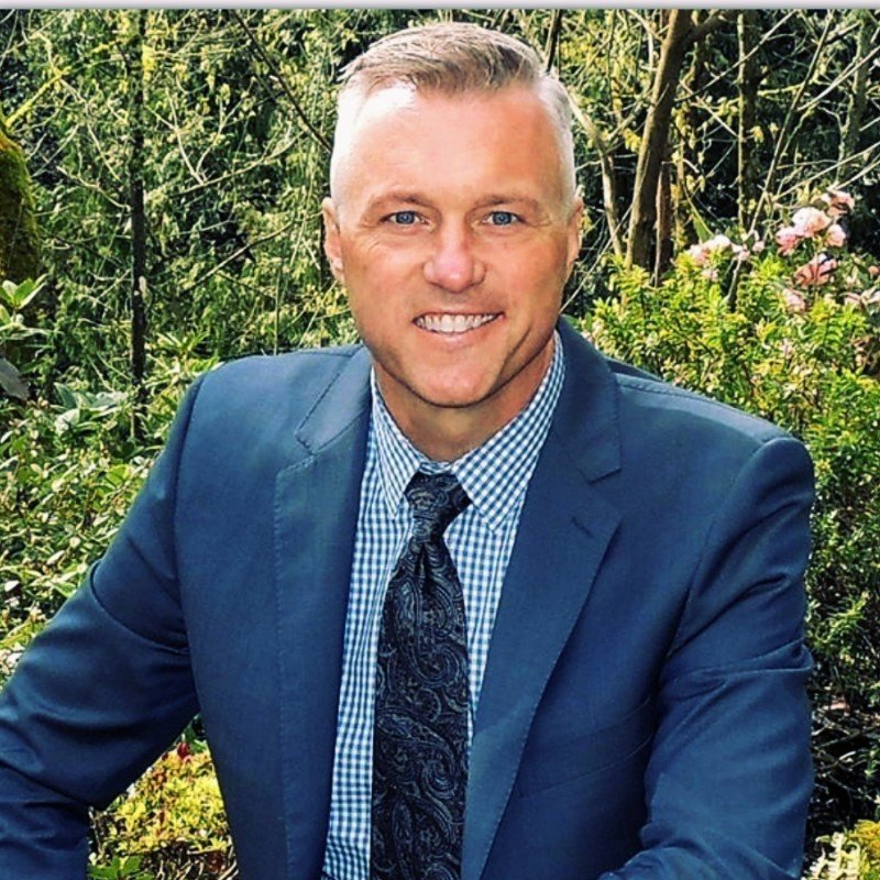A smiling man in a blue suit, blue checkered shirt, and patterned tie, posing outdoors with green foliage and pink flowers in the background.