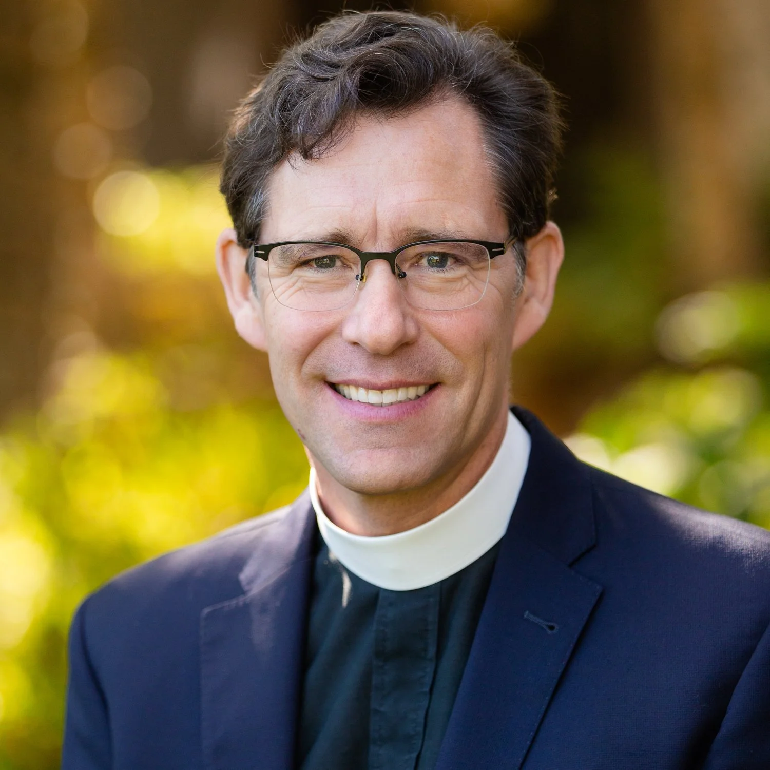 Close-up portrait of a smiling man in clerical attire with glasses, outdoors with blurred greenery in the background.