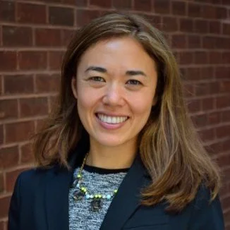 A woman with brown hair smiling in front of a brick wall
