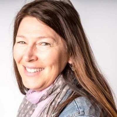 A woman with long brown hair smiling, wearing a denim jacket and a light-colored scarf.