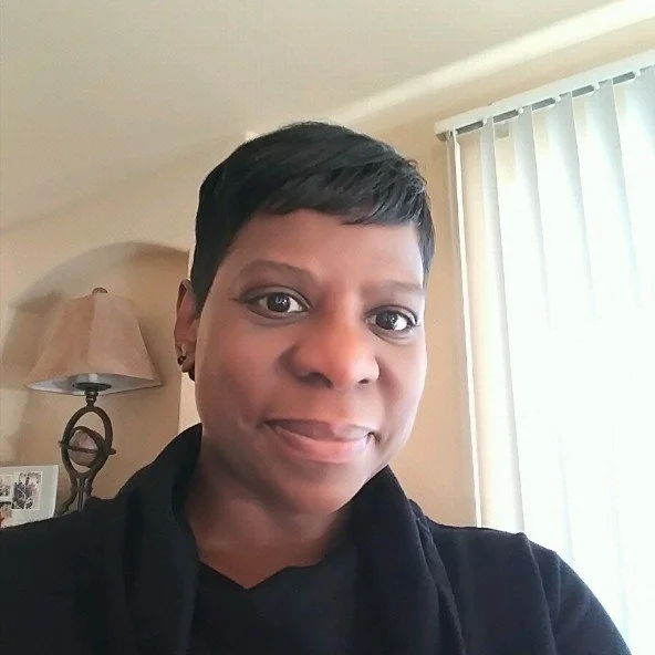 A woman with short black hair and earrings, smiling at the camera, in a room with beige walls, a lamp, and a window with vertical blinds.
