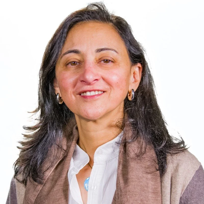 Portrait of a woman with shoulder-length dark hair, smiling, wearing gold hoop earrings, a white shirt, and a multi-colored scarf, against a white background.