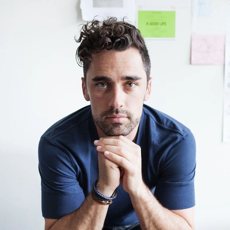 A young man with curly dark hair and a beard, wearing a navy t-shirt, looking seriously at the camera while resting his chin on his folded hands. In the background, a white wall with colorful notes or posters.
