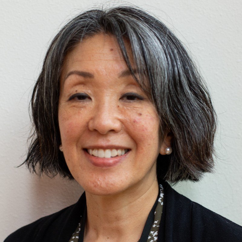 Close-up of an Asian woman with short, wavy black hair, smiling, wearing pearl earrings and a black jacket with a patterned shirt underneath.