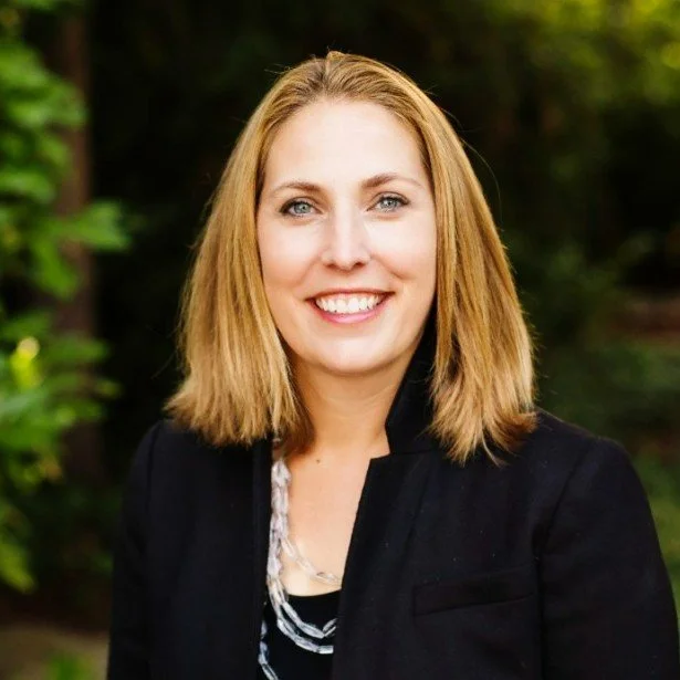 A woman with shoulder-length blonde hair, blue eyes, and a smile, wearing a black blazer and a necklace, standing outdoors with greenery in the background.