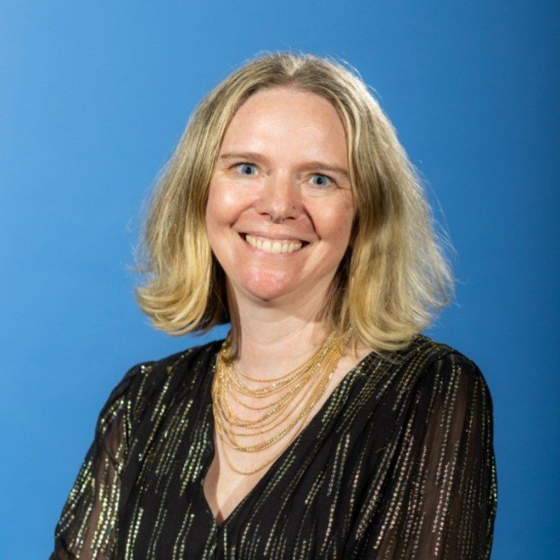 A woman with shoulder-length blonde hair wearing a black, sparkly top and layered gold necklaces, smiling against a blue background.