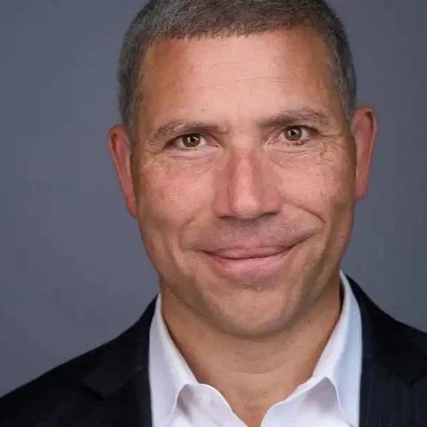 Headshot of a man with short, dark hair, wearing a dark suit jacket and white shirt, smiling at the camera against a gray background.