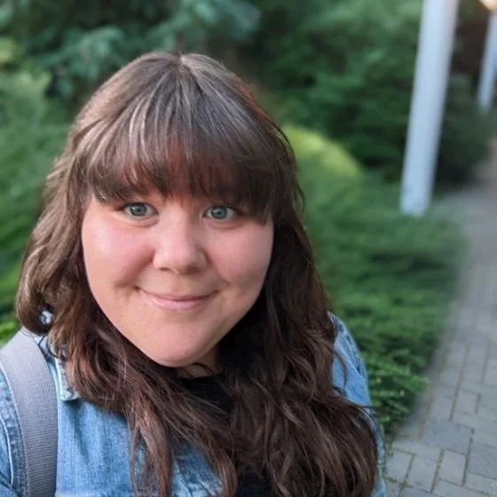 A woman with blue eyes and brown hair with bangs takes a selfie outdoors. She is smiling and wearing a denim jacket.