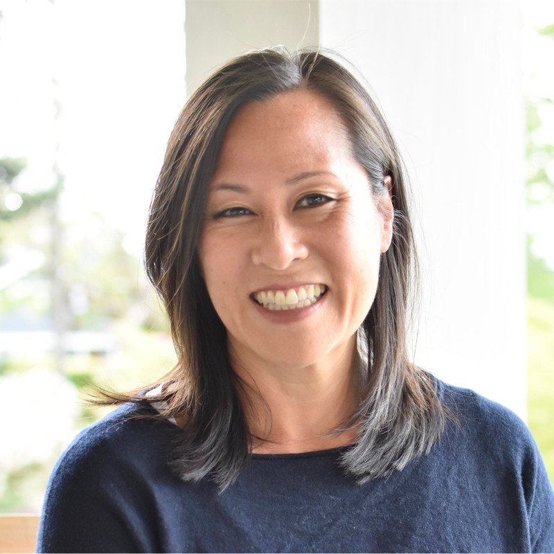 Smiling woman with shoulder-length dark hair, wearing a navy blue top, in a well-lit room with a background of windows showing greenery outside.