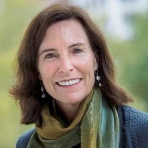 A woman with shoulder-length brown hair smiling outdoors, wearing a dark jacket and a multicolored scarf, with a blurred green and blue background.