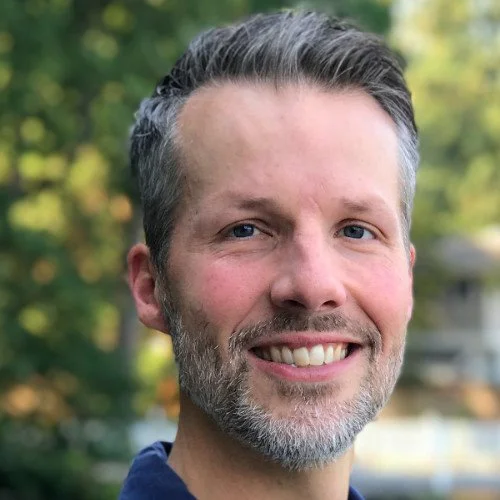 Close-up portrait of a middle-aged man with short gray hair and a gray beard, smiling outdoors with blurred trees and houses in the background.
