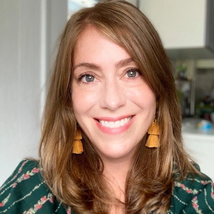 A smiling woman with shoulder-length light brown hair, wearing gold tassel earrings and a dark green floral top.