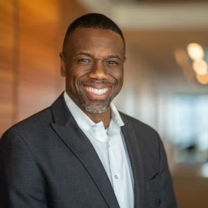 A smiling man in a dark suit with a white shirt, standing indoors.