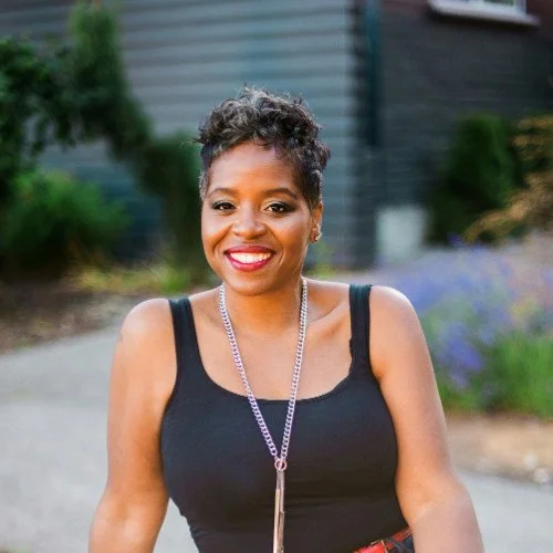 A woman outdoors smiling with short curly hair, wearing a black tank top and a silver necklace.