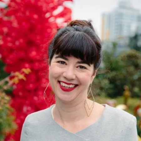 Portrait of a smiling woman with dark hair in a bun, wearing a gray top and dangling earrings, standing outdoors with a red tree and city buildings in the background.