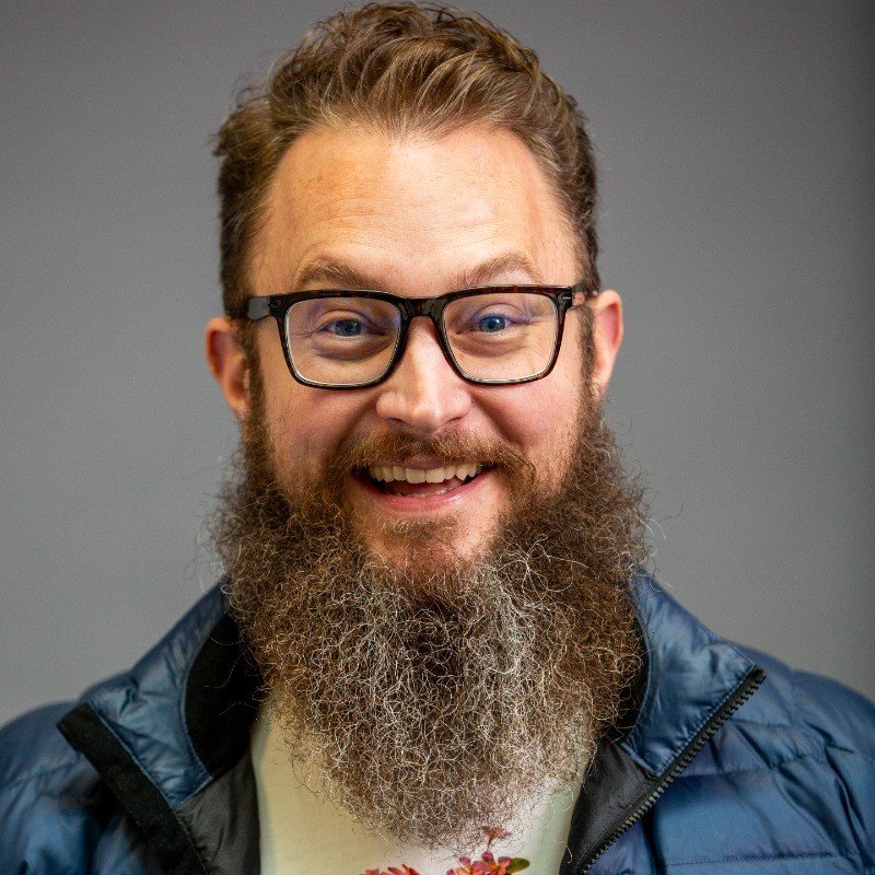 A man with glasses and a full beard smiling, wearing a blue jacket, set against a plain gray background.