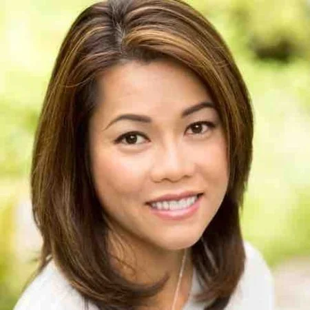 Close-up of a smiling woman with shoulder-length brown hair outdoors with a blurred green background.