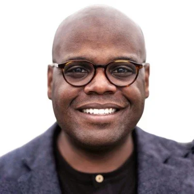 A smiling man with glasses and a shaved head, wearing a dark blazer and black shirt.