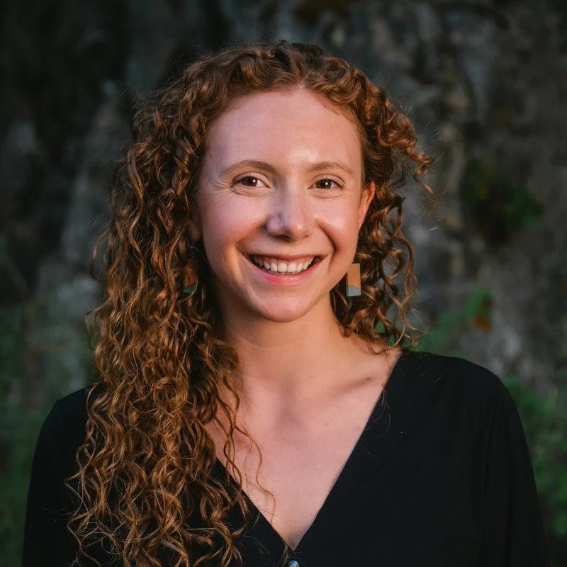 A woman with curly red hair smiling outdoors with a blurred natural background.