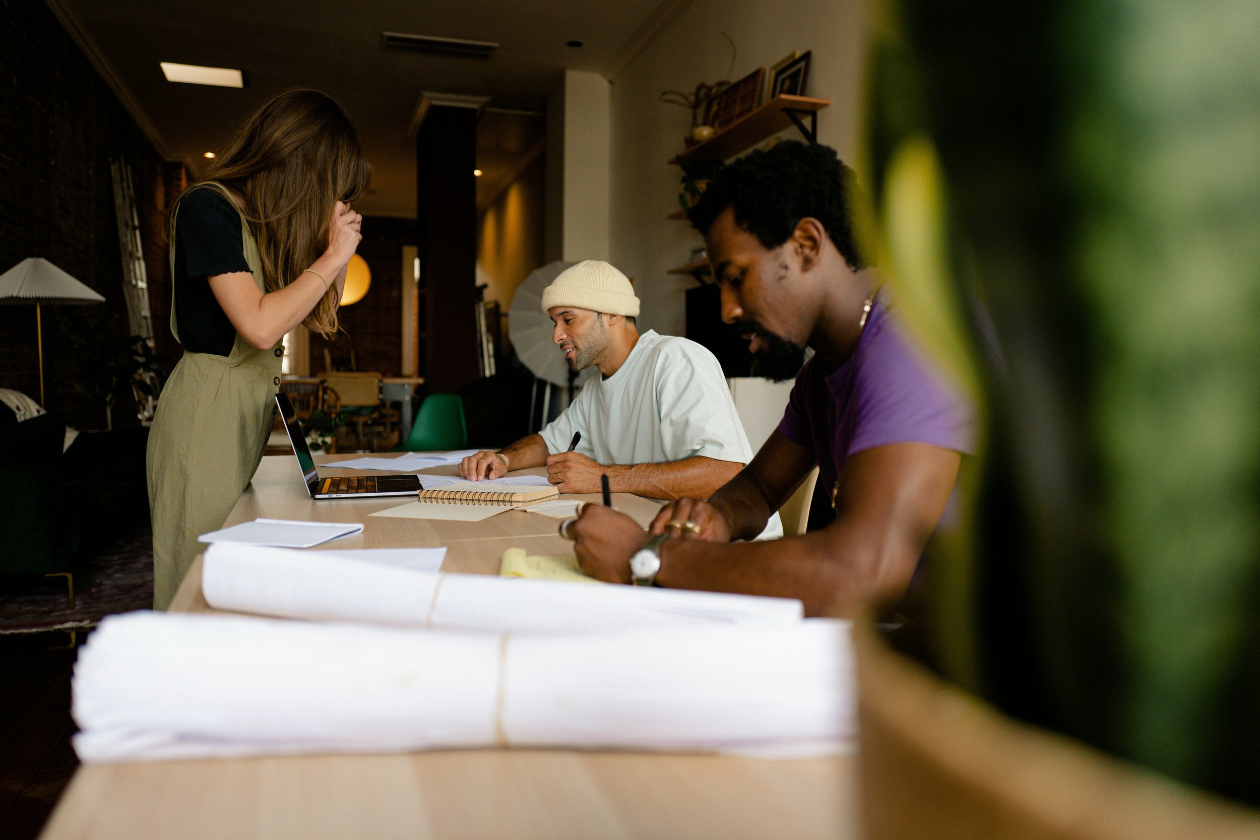 Three people working at a meeting table inside a room, with a woman standing and two men seated, one wearing a beanie and the other in a purple shirt, with papers, notebooks, and a laptop on the table.