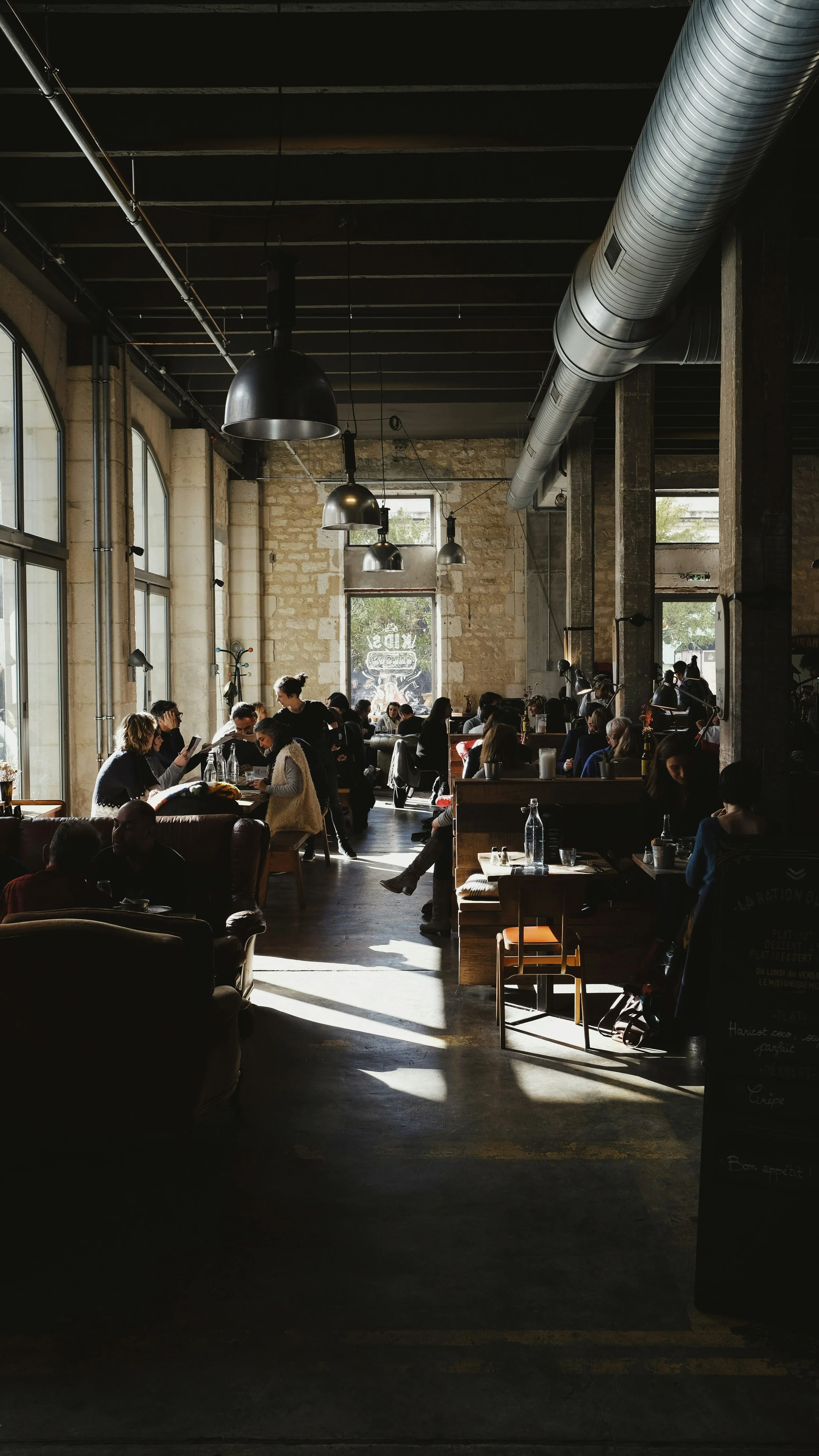 Interior of a busy coffee shop with large windows, high ceilings, hanging lights, and people sitting and chatting.