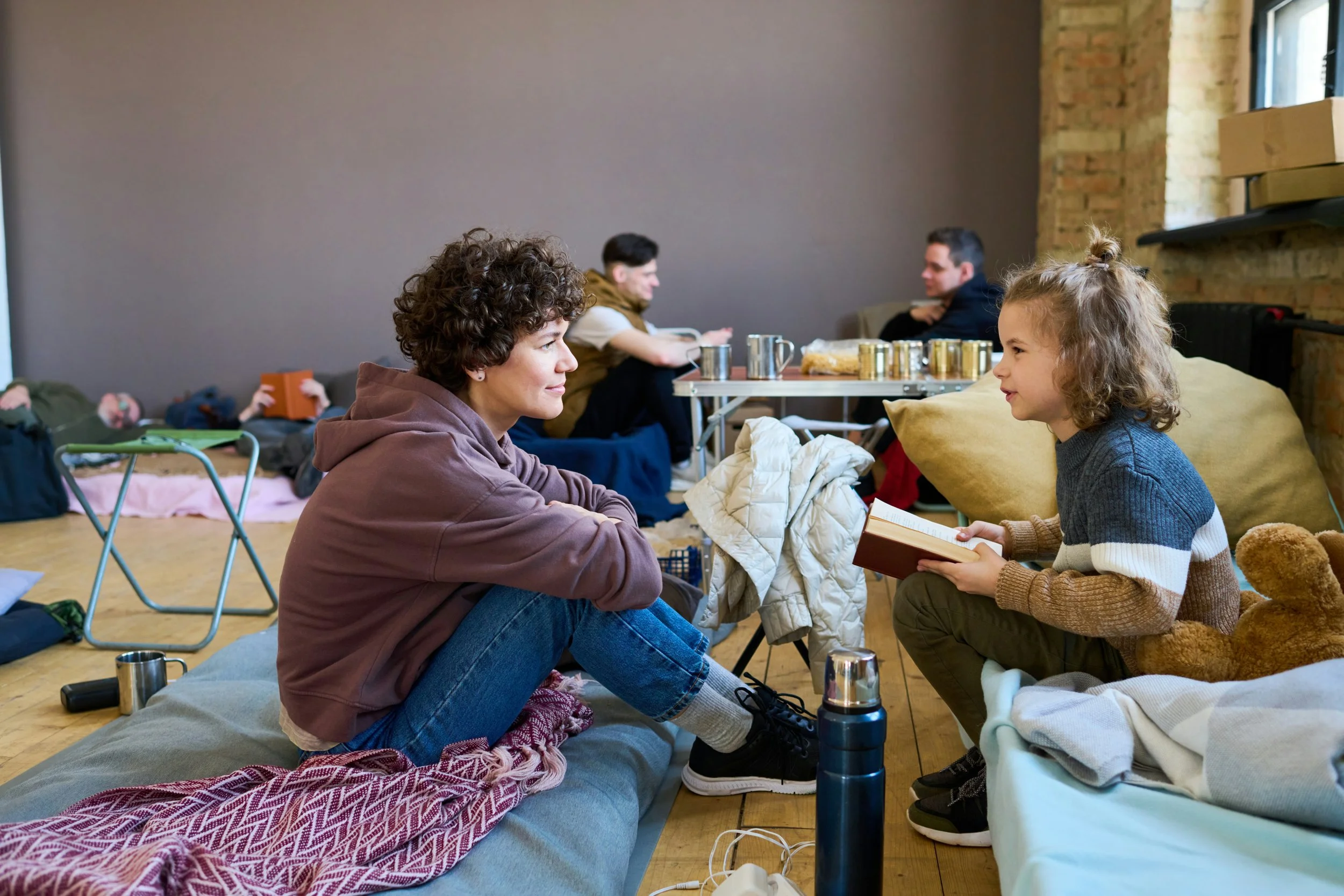 Young woman and girl sitting on the floor facing each other, engaged in conversation, with the girl holding a book. In the background, a group of people are sitting at a table, talking and enjoying breakfast or snacks, in a cozy, casual setting with brick walls and large windows.