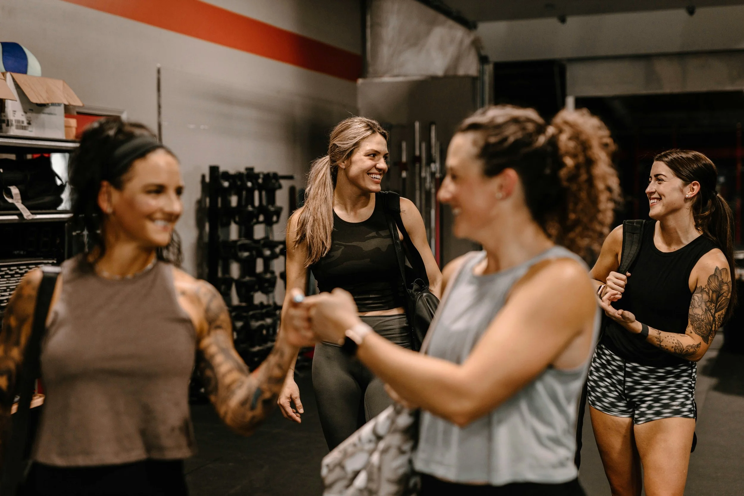 Four women in athletic attire are greeting and smiling at each other inside a gym.