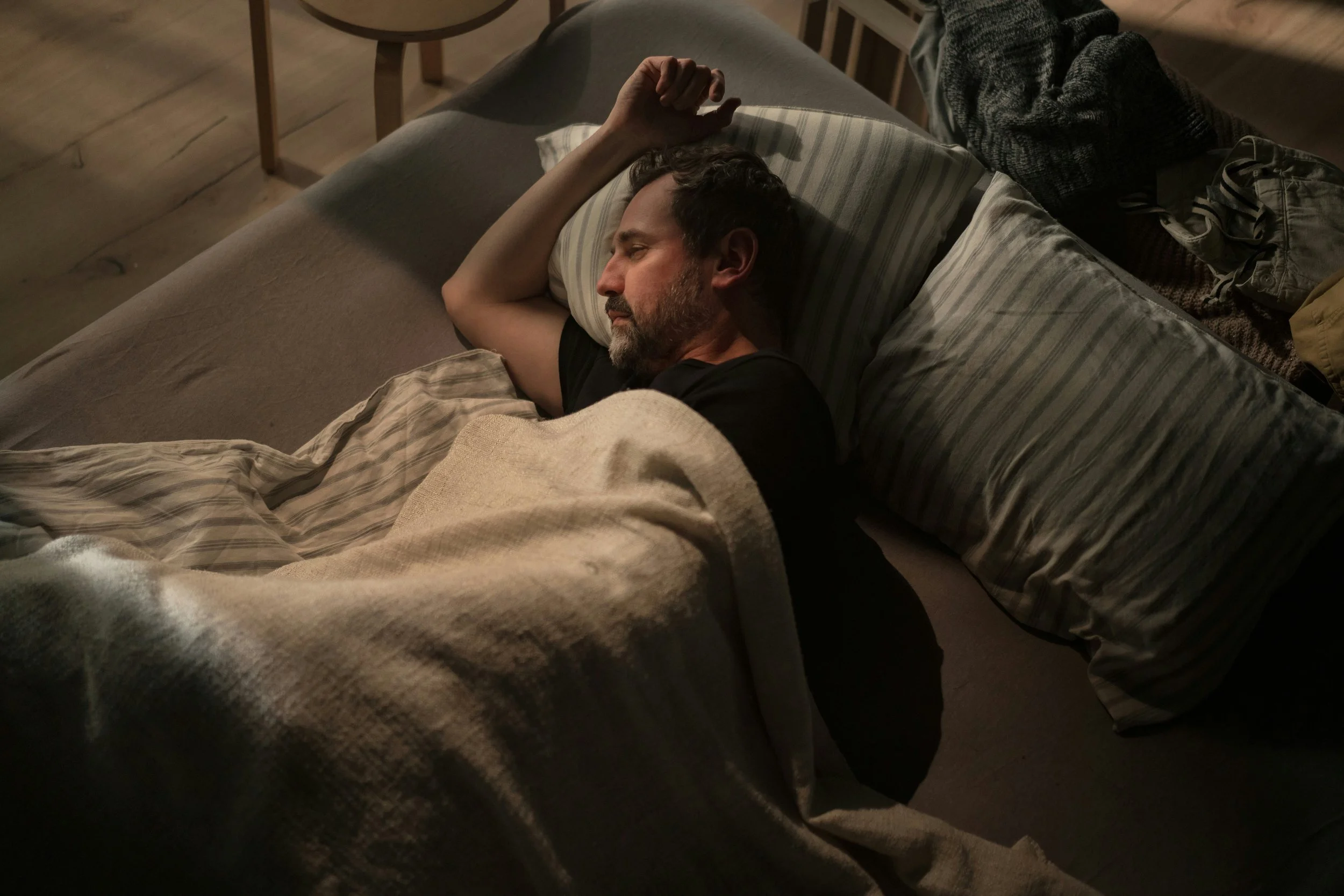 A man with a beard and short dark hair sleeping on his side in bed, covered with a beige blanket, with pillows and clothes around.
